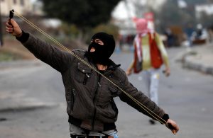 A masked Palestinian demonstrator uses a slingshot to hurl stones towards Israeli troops during clashes at a protest against U.S. President Donald Trump's decision to recognise Jerusalem as the capital of Israel, near the West Bank city
